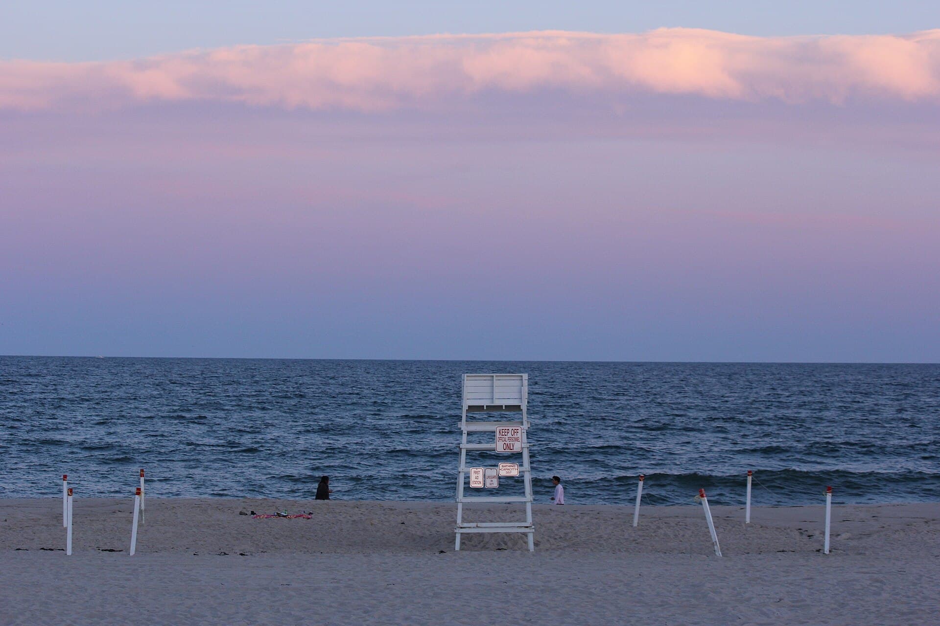Coopers at dusk, with the lifeguard chair empty and its notice board reading Bathing Capacity 357 — the sign that dates the shot to after hours. Calm sea, pink cloud along the horizon, two people on the sand.