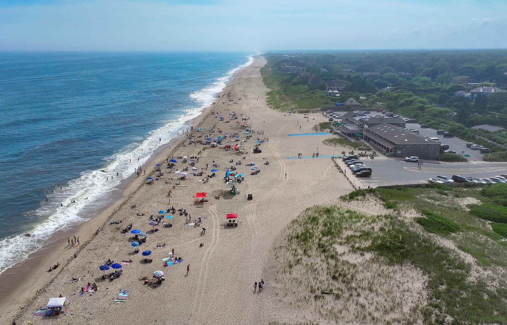 Main Beach from the air on a clear summer day — the shingled pavilion and its car park on the right, umbrellas and swimmers spread along the shore, the ocean running west to Georgica. The pavilion is how you know it is this beach and not another.
