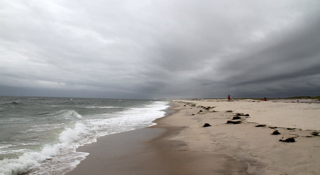 Ponquogue under a heavy dark overcast with a wrack line along the sand and two figures far down the beach. This is a grey unsettled day, not a typical summer afternoon here.