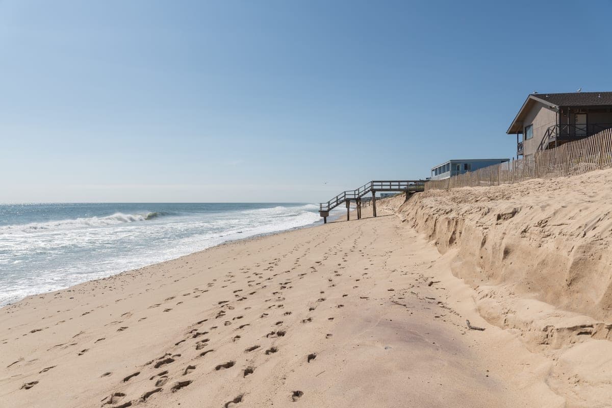 South Edison in early October — empty sand, clear sky, and the houses at the top of the dune with wooden stairs down to the beach. The dune face is visibly cut back by erosion, which is why those stairs end where they do.