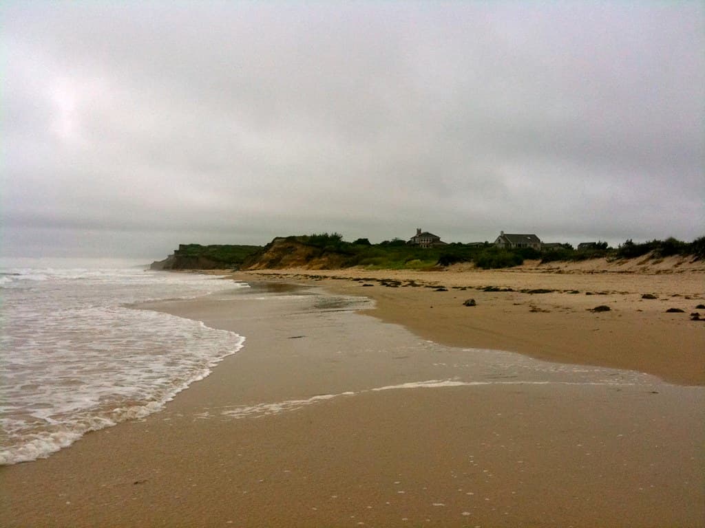 Ditch Plains under flat grey cloud, looking east to the bluff and the houses along its top. The dark clumps along the sand are wrack — seaweed left by the tide, normal here and not pollution.